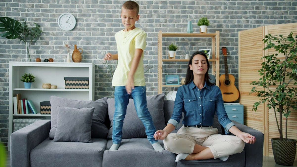 Happy kid boy is dancing on sofa while young mother is meditating in lotus position at home sitting on couch with closed eyes. Childhood and parenthood concept.